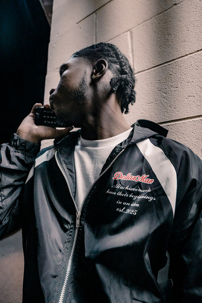 Man in a black and gray jacket with text leaning against a wall.